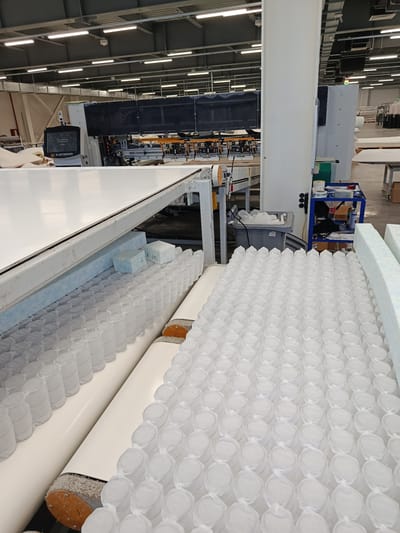 The image captures a wide shot of a mattress manufacturing facility, likely in Zangiata, Uzbekistan, with a focus on the production line. In the foreground, rows upon rows of individually wrapped pocket springs are arranged on a conveyor system, awaiting further processing. To the left, a large, white, flat surface suggests a table or another part of the production machinery. In the mid-ground, a large, industrial machine with a black covering and a control panel displaying the "ELEKTROTEKS" logo is visible, indicating the brand of the equipment. The machine appears to be in operation, with various components and wiring visible. A blue cart laden with supplies, including tape, bottles, and tools, is positioned to the right of the machine. In the background, the ceiling of the factory is lined with fluorescent lights and industrial ventilation systems. The overall environment is a clean, organized, and functional factory floor.