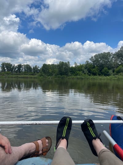 The image depicts a low-angle view of two people's legs and feet from the knees down, sitting in what appears to be a boat on a body of water. The person on the left is wearing brown sandals, while the person on the right is wearing black water shoes with green stripes. Both individuals are positioned on a blue and red inflatable object, likely part of the boat. A white tube or railing runs horizontally across the lower portion of the image.
The background reveals a calm river reflecting a cloudy blue sky and a dense line of green trees and foliage along the far bank, indicating a natural, possibly rural or wilderness setting. The water surface is rippled, suggesting a gentle movement. The scene is set outdoors under daylight, with diffused sunlight filtering through the clouds. The location is identified as the city of Samoradovo, Russia.