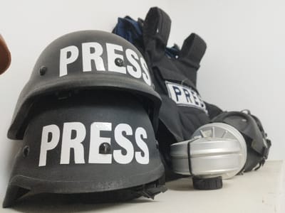 In Dura, Palestinian Territory, a forensic examination of the scene reveals journalistic protective equipment displayed indoors on a light-colored, flat surface against a plain, light wall. In the foreground, two matte black helmets are stacked vertically, each bearing the white, capitalized word "PRESS" on its front. To the right and slightly behind the helmets, a black protective vest is visible, showing a blue and white "PRESS" patch. Adjacent to the vest, a silver cylindrical gas mask filter with a black base rests on the surface, positioned in front of a partially obscured black gas mask or its storage case. No individuals are present in the scene.