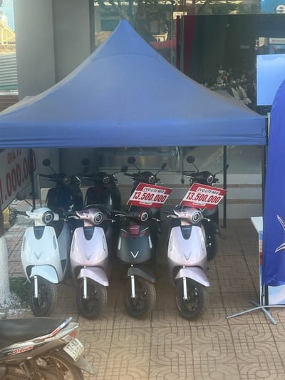 The scene depicts an outdoor commercial display of electric scooters situated on a tiled sidewalk in Trung Hồ, Vietnam. A blue pyramidal canopy tent covers eight VinFast EVO Lite Neo electric scooters arranged in two rows. The front row, from left to right, features one white, two light pink/lavender, and one dark gray/black scooter. Two red signs displaying "EVO LITE NEO 13.500.000" are attached to the handlebars of the two light pink scooters. A partially visible white sign on the far left reads "GIÁ TỪ 1.000.000". In the lower left foreground, a silver and black Yamaha Sirius motorcycle with a partially legible license plate "17-K8 102.XX" is parked on the patterned tiled ground. The background reveals a commercial storefront with large glass windows reflecting ambient light and elements from across the street. A blue vertical banner on a stand is partially visible on the right. No individuals are present in the image. The environment is under bright daylight conditions.