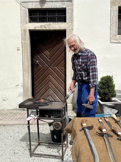 An elderly man with long white hair, wearing a red and black plaid shirt and blue overalls, is standing next to a blacksmith's forge. He is holding a pair of tongs in his left hand and a hammer in his right. The forge is black and has a small motor attached. A small decorative white ribbon is tied to the forge's frame. Behind the man is a white building with a window and a large wooden door with a diamond pattern. To the right of the man is a table covered in burlap, displaying various swords and knives for sale. A small shrub is visible behind the table. The ground is paved with light-colored stones. The scene is taking place in Častá, Slovakia.