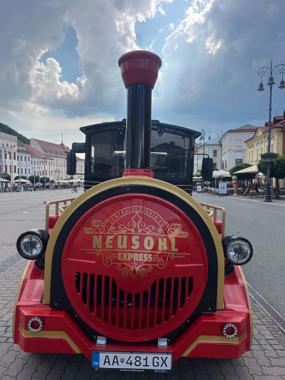 The image shows a close-up view of the front of a red and gold miniature train in an urban square. The train has "NEUSORL EXPRESS" written on its facade in gold lettering, along with "CENTRAL SLOVAKIA.EU" around a decorative emblem. The train has two headlights and a large red smokestack. Behind the train, the square is paved with cobblestones and lined with buildings. Lampposts with decorative wrought iron designs are visible. People are seated at outdoor tables in front of buildings, and some are walking in the background. The sky is partly cloudy with sunlight filtering through. The license plate on the train reads "AA 481 GX" and displays the Slovakian flag. The scene is likely set in the SNP Square in Banská Bystrica, Slovakia, given the context of the train's branding and the architecture.