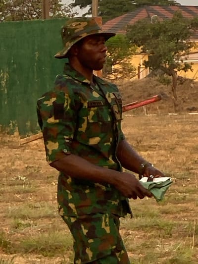 A soldier in camouflage uniform and hat stands outdoors during the day. He looks to his left and holds a folded, green cloth in his hands. Trees, a building, and dry grassy fields surround him. The word "NIGERIA" is visible on his uniform. A thin, red object sticks out from behind his left shoulder. He wears rings and a bracelet on his left hand.