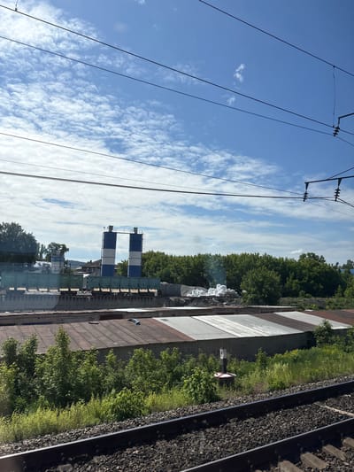 The image captures an exterior view of an industrial area adjacent to a railway in Vyatskiye Polyany, Russia, under a partially cloudy blue sky.

In the upper portion, a bright blue sky is visible, dotted with numerous small, white altocumulus or stratocumulus clouds. Multiple black overhead power lines, part of a railway's catenary system, traverse the sky horizontally, with some angled supports and insulators visible on the far right.

The mid-ground features a landscape dominated by industrial structures and dense greenery. Centrally positioned are two tall, cylindrical silos, each painted with alternating horizontal stripes of blue and white, connected by a narrow overhead walkway. To their left, a smaller, similar silo is partially visible, bearing the text "ELKON." In front of these silos, several green freight wagons are stationary on a raised rail siding or platform. To the right of the main silos, piles of white bags (likely containing dry bulk material such as cement) and grey aggregate are visible on another platform. A faint plume of light-colored smoke or steam emanates from behind these piles on the right side.

Below these industrial elements, a long, single-story building with light-colored brick walls and a multi-paneled corrugated metal roof (featuring sections of rust-brown and grey/silver) extends horizontally across the scene. A blue object, possibly part of a vent or pipe, rests on this roof on the right side. Dense green trees and shrubs form a natural barrier behind the industrial structures and the building, extending to the left and right edges of the frame.

In the lower foreground, two parallel railway tracks are visible, laid on a bed of dark grey gravel ballast. Green grass and smaller shrubs grow between the tracks and the building. A white and red marker post, approximately one meter tall, with the number "6" printed on its upper section, stands upright near the tracks on the right side. No individuals are discernible within the frame. The scene is illuminated by daylight, casting soft shadows.