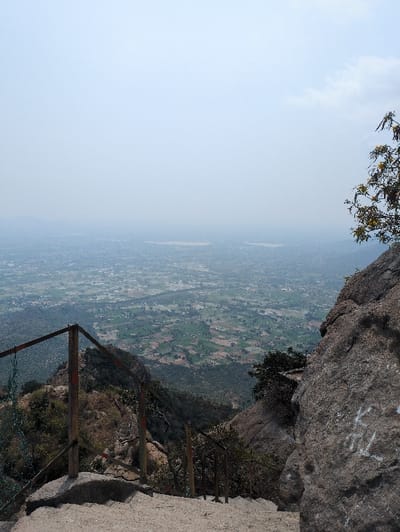 The image displays a high-angle panoramic view looking down from a rocky ascent, presumably within Kadaladi, India. In the immediate foreground, concrete stairs descend towards the left, accompanied by a rusted metal handrail with dark brown posts and a horizontal bar, secured by a green wire mesh on its left side. A large, dark grey rock face occupies the right foreground and midground, bearing white graffiti resembling "KL" towards the center-right. A tree with yellow flowers is partially visible emerging from the top right of this rock face. Beyond the stairs and rock, the terrain drops steeply into a valley characterized by scrub and scattered trees. The expansive background reveals a vast plain featuring a patchwork of green and brown agricultural fields, interspersed with scattered structures. At least two distinct, light-colored bodies of water are visible within these plains. Distant mountains appear on the horizon, obscured by a pervasive atmospheric haze under a pale blue-grey sky. No individuals, specific actions, or interactions are discernible in the scene.
