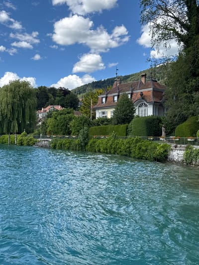 This is a photograph of a lakeside view in Hünibach, Switzerland, featuring a stately home with a red tile roof, a lush green garden, and the turquoise waters of Lake Thun in the foreground. The house, characterized by its dormer windows and ornate architectural details, sits on a gentle slope leading down to the lake. A manicured hedge borders the property, with a stone wall and railing separating it from the water. The lake surface is rippled, reflecting the bright blue sky dotted with fluffy white clouds. Tall trees, including a weeping willow on the left, frame the scene, and a wooded hillside rises in the background.