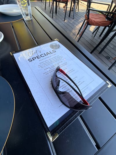 A black outdoor table, likely at a restaurant in Glenview, United States, holds a menu featuring "Chef's Specials." A pair of sunglasses rests on the menu. Tableware is visible, suggesting a meal setting. The table itself is made of dark planks, and chairs with fabric cushions are partially visible, indicating an outdoor dining area. The scene implies someone is about to dine or has just finished a meal.