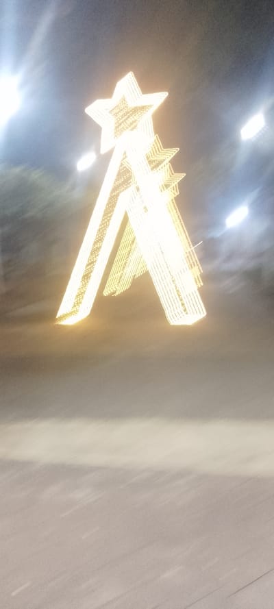 A blurred photograph depicts a structure resembling a stylized Christmas tree, composed of stacked, illuminated stars. The largest star sits atop two angled columns, also lit, creating a triangular shape. These columns are made of parallel lines, presumably light strings. The background is indistinct, with blurred streetlights suggesting an urban setting. The lower portion of the image shows an out-of-focus expanse of pavement or ground. Overall, the image conveys a sense of movement and an out-of-focus capturing.