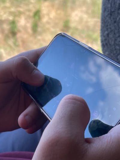 The image shows a close-up of a person's hands holding a smartphone. The phone's screen is reflective, showing a blue sky with clouds and some blurry purple shapes, possibly from a game or app. The screen also has visible cracks. One hand is holding the bottom of the phone, while the other hand's thumb and forefinger are pressing on the side of the device, suggesting interaction with the screen or buttons. The background is blurred, with hints of green and brown foliage and a dark gray textured surface, possibly indicating an outdoor setting or a close-up of a person in a car. The lighting suggests it could be daytime. There is no visible text in the image.