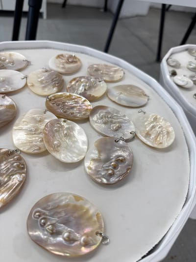 The image shows a close-up view of a white, circular tray filled with oval-shaped pendants made of a pearlescent material, possibly mother-of-pearl. Some pendants have embedded pearl-like embellishments. Each pendant has a small silver-colored loop attached at the top. A second tray of similar pendants is visible in the background. The image was taken in the city of Синьково, Russia.