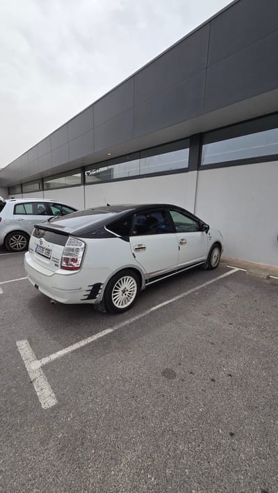 The image shows a white car parked in a parking space next to a modern building. The license plate reads "3336 GBB". The car is parked slightly crooked within the lines of the parking space. The location is indicated to be in Altura, Spain. The building has a gray overhanging structure with windows. The sky is overcast. A second car is partially visible behind the main car.