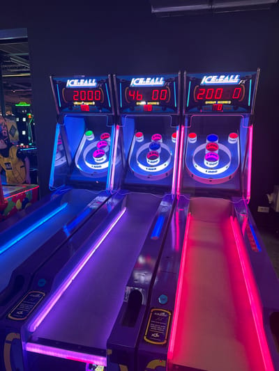Three "Ice Ball FX" arcade games are arranged side-by-side within an entertainment facility located in Saint-André-lez-Lille, France. The environment is dimly lit, with prominent internal LED illumination emanating from the game machines.
The leftmost game displays a red digital score of '2000' and 'YOUR SCORE 0' below it. Its playing lane is bordered by blue LED lights. The middle game displays '46 00' and 'YOUR SCORE 0', with purple-pink LED lighting along its lane. The rightmost game shows '200 80' and 'YOUR SCORE 0', with red LED lighting on its lane. Each game features an inclined ramp leading to a circular target area containing multiple holes designated with point values, including 1,000, 2,000, 3,000, 4,000, 5,000, and 10,000 points. Instructional panels on the lower part of the lanes indicate "PUSH START BUTTON."
A partial view of an individual's torso and left arm is visible on the far left of the frame. The person is wearing a yellow and black sports jersey featuring a 'B&1' emblem. Additional illuminated arcade machines are present in the blurred background to the left.