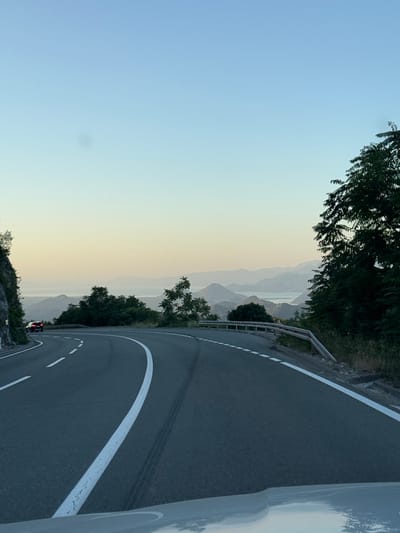 The image shows a winding road with white lane markings, curving to the left. A dark-colored car is visible on the road in the distance, heading in the same direction as the viewer. To the left of the road is a steep, rocky mountainside covered in sparse vegetation. On the right side of the road, there is a guardrail and dense green foliage, including a large tree in the foreground. Beyond the road and vegetation, a panoramic view of rolling, hazy mountains and a body of water can be seen, stretching to the horizon under a clear, pale blue sky transitioning to a soft yellow hue near the horizon, suggesting either dawn or dusk. The overall scene depicts a scenic mountain road in Gornja Brova, Montenegro.