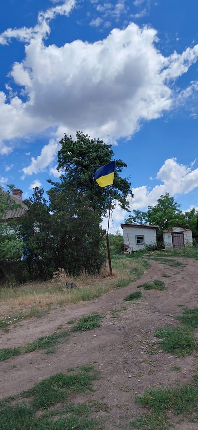 In Ketrysanivka, Ukraine, a Ukrainian flag flies from a pole in front of a large green tree. The flag, divided horizontally into blue and yellow sections, is partially unfurled. To the left of the tree, a house with a grey roof and a red chimney is visible. Beyond the tree, two small buildings stand near a dirt path. One building is white with a single window, and the other is constructed of concrete blocks and has a wooden door. The ground is a mix of dirt and dry grass, with some green vegetation scattered throughout. The sky above is blue with scattered white clouds.