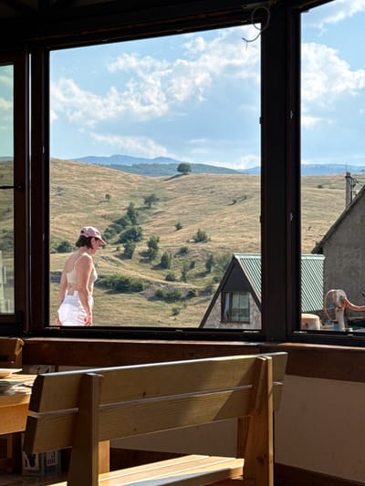The image shows a view through a large window overlooking a hilly, grassy landscape under a partly cloudy sky. In the foreground, inside the building, a wooden bench and a table are visible. Through the window, a woman wearing a pink cap and white shorts is standing and looking out at the landscape. She is wearing a light-colored top. In the middle ground, there is a small, triangular-roofed house with stone and wood accents, and a larger building with a green, striped roof. In the background, rolling hills extend to the horizon, with scattered trees. The overall setting appears to be rural, possibly in Rudinice, Montenegro, given the natural landscape and architecture.