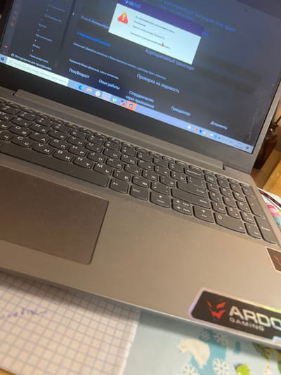The image shows a close-up view of a laptop keyboard and screen, with a portion of a notebook visible in the lower right. The laptop is open, and the screen displays a website with Russian text. The website appears to be a job portal or service, with sections like "Корпоративный транспорт" (Corporate Transport), "Проверка на лидовость" (Verification of Personality), and "Опыт работы" (Work Experience). There is a pop-up window on the screen with a warning icon and Russian text, likely indicating an alert or important message.
On the laptop lid, there is a sticker with the word "VARDO GAMING" and a stylized logo. The keyboard itself is a standard QWERTY layout with some Cyrillic characters mixed in, suggesting it's a bilingual keyboard. The scene appears to be indoors, likely a home or office setting, with a wooden surface visible in the background.
The lighting suggests it might be daytime, but the exact time is unclear. No people are visible in the frame, but the context of the website points to some form of administrative or professional activity. The overall impression is of someone using a laptop for work or research. The presence of the "25.06.2026" date on the screen hints at a future date, possibly related to an event or deadline.