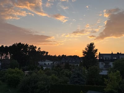 The image depicts a sunset over a suburban area in Marki, Poland. The sky is filled with a gradient of orange, yellow, and pale blue, with scattered clouds catching the warm hues of the setting sun. In the foreground, there is dense foliage, including trees and bushes, with a portion of a dark hedge visible at the bottom. Behind the vegetation, several buildings are visible. These include residential houses with dark tiled roofs and dormer windows, as well as a multi-story apartment building with balconies. A line of tall evergreen trees forms a dark silhouette against the sky in the middle ground. The overall scene conveys a peaceful, residential atmosphere at dusk.