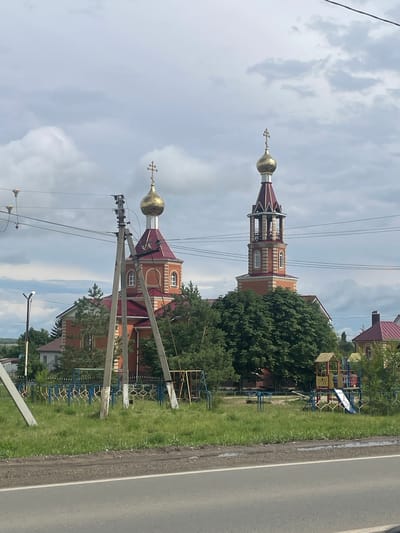The image shows a street scene in a rural area, likely in Дубки, Russia, on a cloudy day. The main subjects are two domed churches with red roofs and golden crosses. One church is taller and has a bell tower. There are power lines running across the foreground, obscuring parts of the scene. A grassy area with a playground is visible to the right of the churches, bordering a paved road. The overall impression is that of a quiet, possibly residential, village setting. There is no visible text in the image.