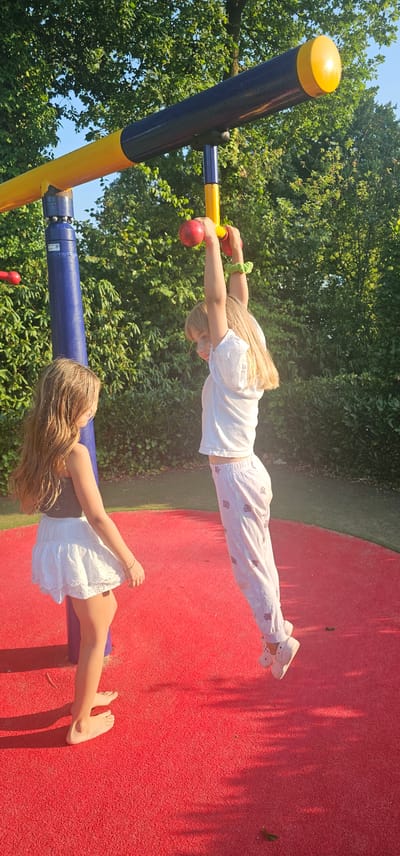 Two young girls are playing on a playground in Groenlo, Netherlands. The girl on the right is hanging from a yellow bar with red handles, her arms outstretched. She is wearing a white shirt and white pants with a subtle pink pattern. The girl on the left is standing and looking up at the other girl. She is wearing a white skirt and a brown top. They are on a red, rubberized surface. The background consists of green trees and a blue sky. The sun is casting shadows on the ground.