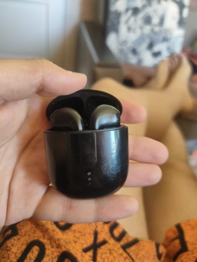 A close-up shot shows a person's hand holding a black charging case for wireless earbuds. The case is open, revealing two black earbuds inside. The charging case has three small circular indicators on its front. The background is blurred, but appears to be an indoor setting with a person's legs visible, possibly lounging. The image was taken on August 12, 2023, in Salavat, Russia.
