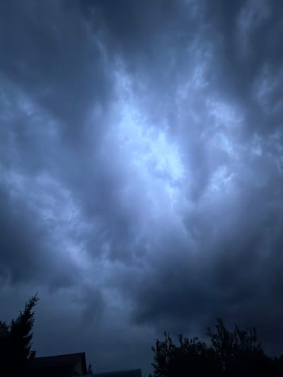 The image captures a dark, overcast sky filled with thick, blue-grey storm clouds. A faint, bright area of lighter cloud is visible in the upper center of the frame, suggesting a break or thinning of the clouds. The bottom of the image shows the silhouettes of trees and the roofline of a building against the darkened sky. The overall impression is of an impending storm. The location is identified as Манихино Снт, Russia, though no specific landmarks or details within the city are visible to confirm this. There are no people or visible actions taking place. The environment is characterized by dramatic cloud formations and limited visibility due to the darkness.