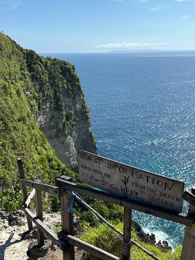 Captured at Bunga Mekar, Indonesia, the image presents an elevated perspective of a cliffside meeting the ocean. Lush vegetation blankets the cliff, transitioning into rocky terrain at its edge. The ocean stretches towards the horizon, displaying varying shades of blue. A weathered wooden sign with the inscription "TOURIST INFORMATION" stands prominently, providing information such as "OPEN FROM TOP 07:00AM~17:00PM" and "FROM THE BEACH CLOSED." The sign is supported by a basic wooden structure with a blue rope detail, set against the backdrop of the cliff and the ocean.