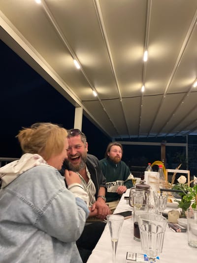 A group of people are gathered at a table on an outdoor terrace. A man with a beard is laughing and embracing a woman with blonde hair. Another man, wearing a green sweater, is looking up. The table is set with glasses, bottles, and other items. The terrace is covered by a roof with built-in lighting. The location is Trebesin, Montenegro.