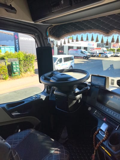 The image captures the interior of a truck cabin, looking outwards through the windshield. The driver's seat, upholstered in black diamond-stitched leather, is partially visible in the foreground. The dashboard features a steering wheel, a GPS navigation device, and various control buttons. Hanging from the top of the windshield are decorative tassels. Outside, a parking lot is visible with several vehicles, including vans and cars. A building with signage reading "GROUPE SIMON PROFIL+" is also present. The scene appears to be set in an industrial or commercial area. The location is identified as Saint-Herblain, France, near a PROFIL+ establishment, likely a tire and auto service center. No people are directly visible in the cabin or the immediate outdoor area, suggesting a moment of pause or an unattended vehicle. The overall lighting indicates daytime with bright sunlight.