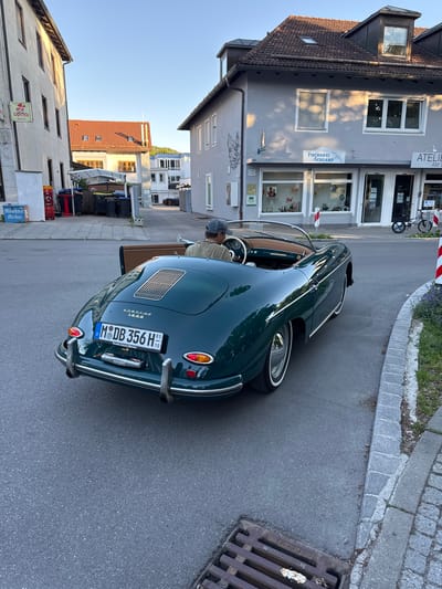 The scene captures a green Porsche 356 B parked on the street in Herrsching am Ammersee, Germany. A man is seated in the driver's seat. The vehicle's license plate reads "M DB 356 H". The shot includes street details, buildings, signage, a storefront labeled "FISCHEREI SCHLAMP," and an "ATELIE" sign. Other objects include trash cans, bicycles, and utility markings on the road. The location is suggested by text on the numberplate.