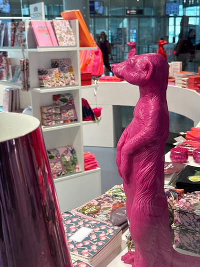 The image shows a display of stationery and decorative items in a retail environment. In the foreground, a bright pink, stylized sculpture of a meerkat stands prominently. The sculpture has a textured surface resembling fur and is posed upright, with its forelimbs held together. To the left of the sculpture, a large, reflective metallic purple cylinder is partially visible, also likely a decorative object or container.
Behind the meerkat sculpture, shelves are filled with various paper products, including notebooks, journals, and possibly patterned pouches. The designs on these items feature floral motifs and flamingo patterns, rendered in vibrant colors. Some items are stacked neatly, while others are displayed upright.
In the background, through a glass partition, a blurred view of a public indoor space can be seen. There are reflections on the glass, and the presence of other shoppers and displays suggests a store or a shopping mall. The architecture visible in the background includes ceiling lights and potentially shop signage. The overall impression is one of a modern retail setting with a focus on colorful and decorative merchandise. Based on the context of the store "Cheddon", this scene is located in the city of Freising, Germany.