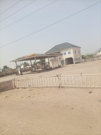 The image depicts an outdoor scene in Gulum, Nigeria, under a hazy, clear sky. In the midground, a gas station operates under a rectangular canopy with "UMARU SALEM WORLDWIDE LTD" inscribed on its fascia. Multiple fuel pumps are visible beneath the canopy. Adjacent to the gas station is a two-story building with a light-colored facade and a dark-tiled roof. The building features multiple blue-framed windows on the upper floor and what appear to be garage or store entrances on the ground floor. The immediate foreground consists of dry, reddish-brown earth with sparse vegetation and debris. A light-colored metal barrier, equipped with wheels and spiked protrusions on its upper edge, stretches across the foreground. No individuals are present, and no active interactions are observed. A single dark-colored flip-flop or sandal lies on the ground near the metal barrier. Distant hills are faintly visible in the background, and overhead power lines traverse the sky.