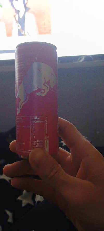 A person's hand is holding a pink Red Bull can. The can has nutritional information and the Red Bull logo printed on it. In the background, a television screen displays an anime character. The scene appears to be indoors, possibly in a living room. The image does not provide information about the specific location within Lipí, Czechia.