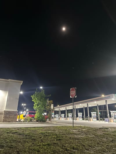 At night in Homosassa Springs, Florida, a well-lit 7-Eleven gas station is visible. Gas pumps are under a brightly lit awning. A stop sign stands near the pumps. A dark sky, with a bright moon, contrasts with the lit environment. Grass lines the right side of the scene. A partially visible building with exterior lighting and landscaping is on the left. People appear to be present, though details are indistinct. A red vehicle is parked beyond the gas station.