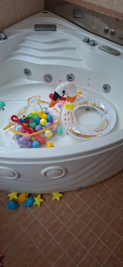 A high-angle, close-up shot shows a white, rectangular bathtub filled with various toys. A clear inflatable ring with gold glitter and an orange and white flamingo design rests in the tub. Next to it is a clear plastic bag filled with colorful plastic balls, a red toy rake, and a blue and green toy. Small, colorful plastic sea creatures, including turtles and fish, are scattered around the inside of the tub. On the tiled floor surrounding the tub, several yellow star-shaped toys and blue droplet-shaped toys are visible. The scene appears to be indoors, likely a bathroom. The lighting is bright, suggesting daytime. No people are visible, and no text is present in the image.