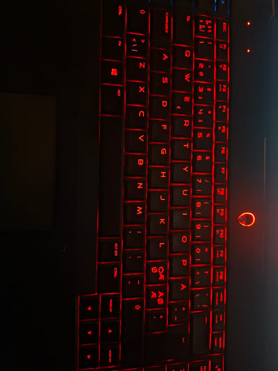 The image shows a close-up of a black gaming keyboard with red backlighting. The keyboard is oriented vertically, with the top edge of the image showing the F keys and the bottom edge showing the arrow keys and modifier keys like Ctrl and Alt. The keys are illuminated with a bright red glow, highlighting the letters and symbols on each key. In the center right of the image, a prominent circular logo with a red glow, consistent with an Alienware logo, is visible on the laptop casing adjacent to the keyboard. The overall scene is dark, with the red light from the keyboard and logo being the primary sources of illumination. The image's orientation and lack of surrounding environmental context suggest it is a product shot or a detail shot of the keyboard itself, rather than a scene captured in a specific urban location like Užuežerė, Lithuania.