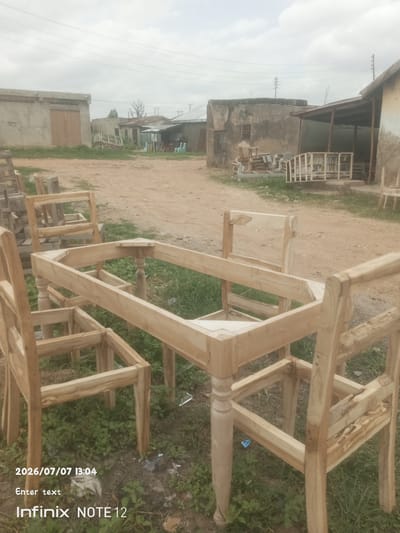 The image shows an unfinished wooden dining set, likely a table and chairs, placed outdoors in what appears to be a workshop or yard. The furniture is made of light-colored wood and is in various stages of construction. The table has turned legs and an octagonal frame. The chairs have simple backrests and legs. The scene is set against a backdrop of several buildings, some appearing to be under construction or in disrepair, with a dusty ground and sparse vegetation. The sky is overcast, suggesting a cloudy day. In the distance, power lines are visible. The overall impression is one of a rustic, working environment. Visible text at the bottom of the image indicates the date and time as "2026/07/07 13:04" and the phone model "Infinix NOTE 12". There is also placeholder text "Enter text". The location context of Kinkinau, Nigeria, suggests an African setting.