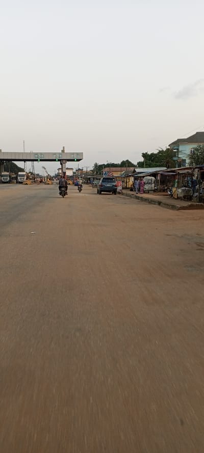 A wide, dirt road stretches into the distance, passing beneath a toll booth structure with green lights illuminated. Motorcycles and a dark-colored car are present on the road. The roadside features open-air stalls and small shops. In the background, buildings and trees are visible under a hazy, light sky. The road appears to be the primary focus of the image, suggesting a journey or passage.