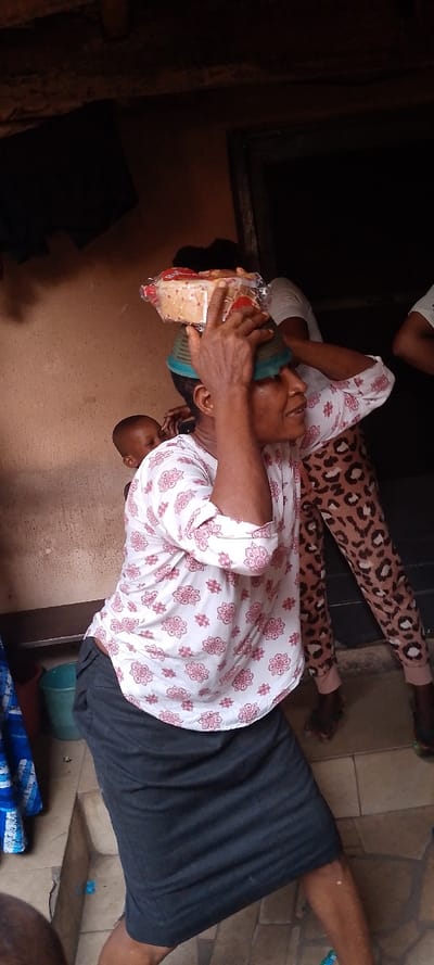 The image depicts a scene in Ohafia, Nigeria, showing a woman in the foreground carrying items on her head and a child on her back.
The primary subject, a woman with dark skin, is positioned slightly off-center to the left, facing right. She wears a white long-sleeved shirt with red floral patterns and a dark grey skirt. On her head, she balances a teal-colored bowl, which supports a clear plastic-wrapped package of what appears to be sliced bread with red accents. Her right hand is raised, fingertips lightly touching the bread package, presumably for stability. Her mouth is slightly open, and she looks towards the right side of the frame. A child's head and shoulder are visible on her back, to the viewer's left.
In the mid-ground, behind the primary subject, partial views of at least two other individuals are discernible. One person wears light-colored pants with a dark brown leopard print pattern. The background consists of a light brown, textured wall. To the right, a dark, recessed doorway or opening is visible. The upper portion of the frame reveals a dark, somewhat uneven ceiling structure, possibly with exposed wooden beams. The lower portion shows a tiled floor with light-colored, rectangular tiles. To the far left, a blue bucket and a partial red object are visible on the floor.