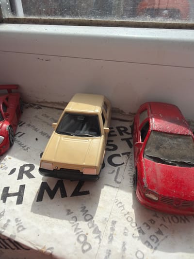 The image depicts three miniature model cars positioned on a printed surface, likely paper or cardboard, which rests on a dirty white window sill. Above the sill, a grimy window pane and its white frame are visible.
From left to right, the visible subjects are:
1. A partial view of a red, low-slung sports car model, appearing in relatively good condition, with a prominent rear spoiler.
2. A full view of a beige hatchback model, identified as a Skoda Felicia, with black trim and dark windows. This model appears well-preserved.
3. A full view of a red hatchback model, identified as a Volkswagen Golf Mk4, showing significant wear, paint chipping, dirt, and damaged or missing windows.
The printed surface beneath the cars displays fragments of English text, including words and letters such as "OM TH," "M Z," "DOING," "HARBOR," "VILLAGE," "ARE," "CT," and "LL NEW," oriented at various angles. The window sill and window pane are visibly dirty and scuffed. Natural light enters from the left, casting distinct shadows of the models onto the surface.
The scene is an interior setting. While the prompt mentions Kermen, Bulgaria, there is no visual information within the image to confirm this specific geographical location. The environment is an indoor window area. No people are present.