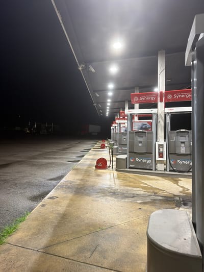 The image depicts an outdoor scene at night, likely a gas station, in Wildwood, United States. A row of illuminated gas pumps is visible under a covered structure with overhead lighting. The ground appears wet, with reflections of the lights. In the background, a semi-trailer truck is parked in the darkness. The overall impression is one of a quiet, possibly deserted, service area.