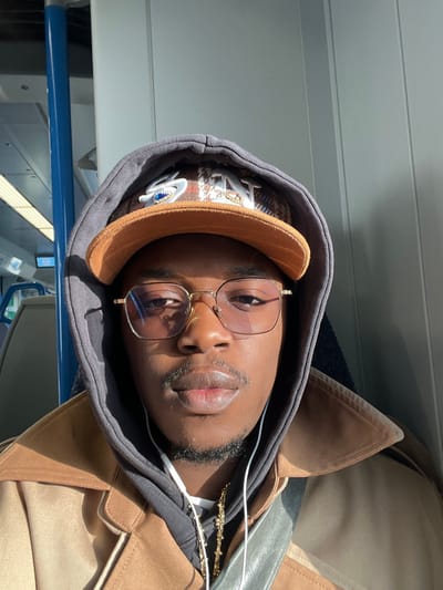 A single individual, a dark-skinned male with a light mustache and goatee, is depicted in a close-up, front-facing selfie taken inside a train carriage. He wears gold-rimmed rectangular glasses with light-tinted lenses, a dark grey hooded sweatshirt with the hood up, and a tan/brown trench coat. Under the hood and over the sweatshirt, he wears a brown corduroy baseball cap adorned with a stylized eye patch featuring a blue gem and another patch spelling "HAT." White wired earphones are visible around his neck, along with a gold-colored chain. A grey strap, likely from a bag, crosses his chest. Strong natural light illuminates the right side of his face (viewer's right), casting a shadow on the left and reflecting off his glasses. The background reveals typical train interior elements: a vertical blue grab pole on the left and a light grey/white ribbed panel wall behind him. The scene is consistent with being on a train operating within or passing through the city of Stevenage, United Kingdom.