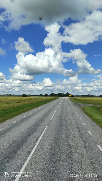 A paved road stretches into the distance under a bright blue sky dotted with dramatic white and gray clouds. The road is flanked by fields of green and yellow crops, with a line of trees visible on the horizon. A yellow sign is posted on the right side of the road in the distance. The timestamp in the lower right corner indicates the image was taken on July 10, 2026, at 3:06 PM. The lower left corner displays "REDMI NOTE 9 PRO" and "AI QUAD CAMERA," indicating the device used for the photograph and its camera features. This scene is consistent with the rural landscape of Sagevere, Estonia, known for its agricultural fields and open roads.