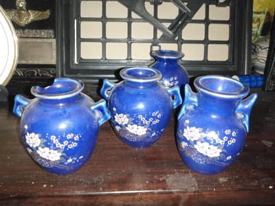 The image depicts an indoor scene in Steenbergen, Netherlands, showcasing four small, glossy, deep blue ceramic vessels on a dark, dusty wooden surface. Three identical vessels in the foreground are ovoid with two small handles and white floral patterns; the leftmost one has a chipped rim. A fourth, smaller, handle-less blue vessel is visible behind the central foreground item. The background includes a partial white plate with a gold rim, a dark plaque reading "ASSUS" with a metallic winged insignia, and a large dark-framed object featuring a grid pattern. Additional indistinct decorative objects are partially visible to the far right. No individuals or active interactions are present.