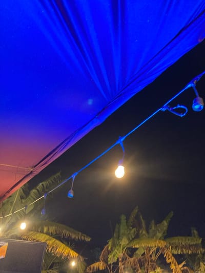 A low-angle nighttime photograph depicts an overhead scene within Murera ward, Kenya. The upper portion of the frame is dominated by a dark blue fabric canopy or tarp, displaying visible folds and some surface speckling, with a reddish-purple hue discernible along its lower-left edge. A string of suspended globe-style lights extends diagonally from the upper-right towards the lower-left. Several bulbs are visible, one prominently illuminated and emitting a warm yellow glow, while others appear unlit or cast a faint blue-white light. Below the string lights, the silhouettes of multiple tropical trees, characterized by their broad fronds and dark foliage, are visible against a black, starless night sky. A dark, rectangular object, possibly a speaker or sign, is partially visible in the lower-left quadrant. No individuals, specific actions, or interactions are discernible.