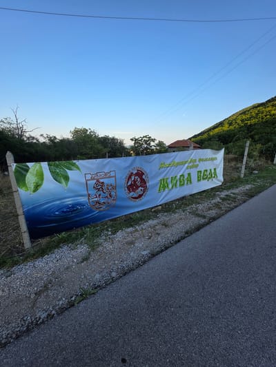 This media file captures a banner with Cyrillic text and imagery, displayed outdoors. The banner advertises a "Международен фолклорен събор" (International Folklore Festival) and promotes "АКИВА ВОДА" (Akiva Water). The text also includes "ДИМОВО" (Dimovo), likely a location or organization name, and "ПАИСИЙ ХИЛЕНДАРСКИ-1927" which refers to a historical figure and a founding year.

The banner itself is the primary object, featuring a stylized emblem with a lion-like creature and water ripples, along with images of dewy green leaves. It is attached to poles and appears to be situated by the side of a paved road, with a gravelly shoulder. In the background, there are trees, some greenery, and a glimpse of a building with a red roof. The scene is illuminated by natural light, suggesting daytime, with a clear blue sky visible. The orientation of the image is somewhat tilted. The overall context is that of an outdoor event or advertisement, likely in or near Dimovo, Bulgaria, given the place name and the presence of a road and natural surroundings.