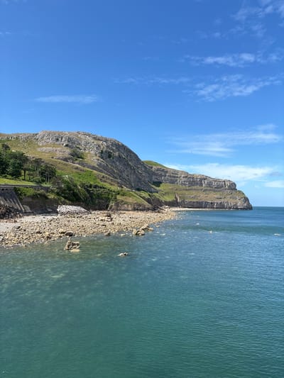 The image captures a scenic view of the Great Orme, a headland on the north coast of Wales, near the town of Llandudno in the United Kingdom. A tall, limestone cliff dominates the center of the image, its face marked by horizontal striations and a patchwork of green vegetation clinging to its slopes. At the base of the cliff, a narrow stretch of rocky coastline transitions into the sea. The water is a pale turquoise color, clear enough to reveal the submerged rocks beneath its surface. The sky is a clear blue with wispy white clouds scattered across it. The image is taken from a low angle, emphasizing the height of the cliff and the expanse of the sea.