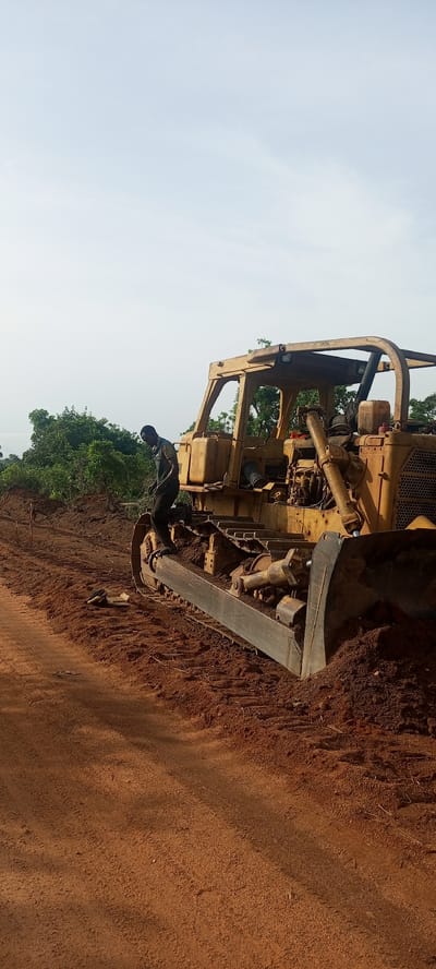 This image captures a scene of land development or construction in Otunade, Nigeria, featuring a large bulldozer and a worker under a clear sky.

The central subject is a robust, yellow crawler bulldozer, appearing well-used with visible wear and tear. It is positioned on a reddish-brown earth surface, characteristic of lateritic soil often found in tropical regions. The bulldozer's large blade is at the front, partially pushed into a mound of earth, suggesting it has either just completed digging or is about to commence operations. Its heavy-duty tracks are also covered in the same reddish-brown soil.

A person, dressed in dark work attire, is standing on the left-side track assembly of the bulldozer. They appear to be focused on the machinery, possibly engaged in an inspection, maintenance, or preparing for operation. Their posture indicates active engagement with the equipment.

The setting is an outdoor, possibly rural or semi-rural landscape. Behind the bulldozer, a dense line of vibrant green trees and bushes stretches across the mid-ground, providing a natural backdrop to the industrious scene. The sky above is a pale, clear blue with very few clouds, indicating bright daylight conditions, likely morning or late afternoon, given the soft illumination without harsh shadows.

The foreground is dominated by the reddish-brown earth, which forms a path or cleared area extending towards the left of the frame. The overall impression is one of ongoing work in a natural environment, utilizing heavy machinery for earthmoving or land preparation. No discernible text is clearly visible on the equipment or in the environment.