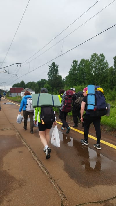 The image shows a group of people walking on a wet, paved surface next to train tracks under an overcast sky. The people are wearing backpacks and carrying bags, suggesting they are travelers or hikers. The environment appears to be outdoors, with trees and buildings visible in the background. The location is identified as Orekhovo, Russia. The pavement has yellow markings along the edge.