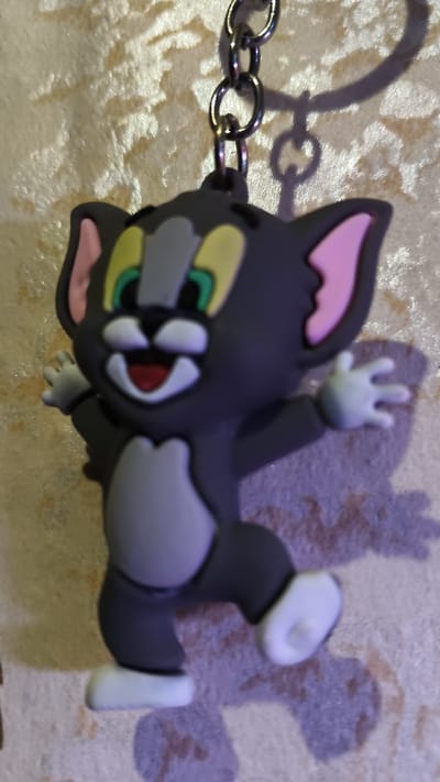The image shows a dark gray keychain shaped like the cartoon character Tom Cat. Tom is depicted with a wide, smiling mouth, bright yellow eyes with green pupils, and pink inner ears. He is shown in a dynamic pose, with one arm raised and outstretched, and his legs spread apart as if in mid-stride or dance. His belly and muzzle are light gray, and he has white paws and feet. The keychain is attached to a metallic chain.

The background is a textured surface, possibly a wall or fabric, with a rough, light-colored texture. Shadows are cast on this background, suggesting a light source is present, though the time of day and weather conditions are not discernible. The shadows also give a sense of depth to the image. The focus is clearly on the Tom Cat keychain, with the background slightly out of focus. No text is visible in the image. The context provided indicates the location is Mokshan, Russia, but there are no visual cues within the image itself to confirm this.