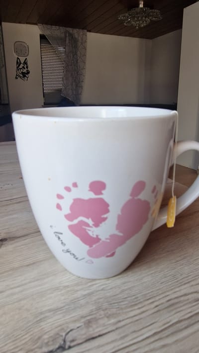 This is a media file of a white mug with pink handprints and the text "i love you!" printed on it. The mug has a tea bag tag attached to its handle. The mug is placed on a wooden surface, which appears to be a table or counter. In the background, there is a glimpse of a kitchen appliance and a decorative element resembling a chandelier. There is also a sticker of a dog and a clock on a white surface. The image is taken indoors, and the lighting suggests it could be daytime. The overall impression is that of a personal and sentimental item, possibly a gift. The location context is Villingen-Schwenningen, Germany, but there are no specific visual cues in the image to confirm this.